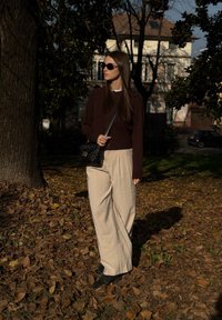 En person står på et tæppe af efterårsblade, iført en mørkebrun sweater, beige bukser med vide ben og holder en sort quiltet håndtaske.