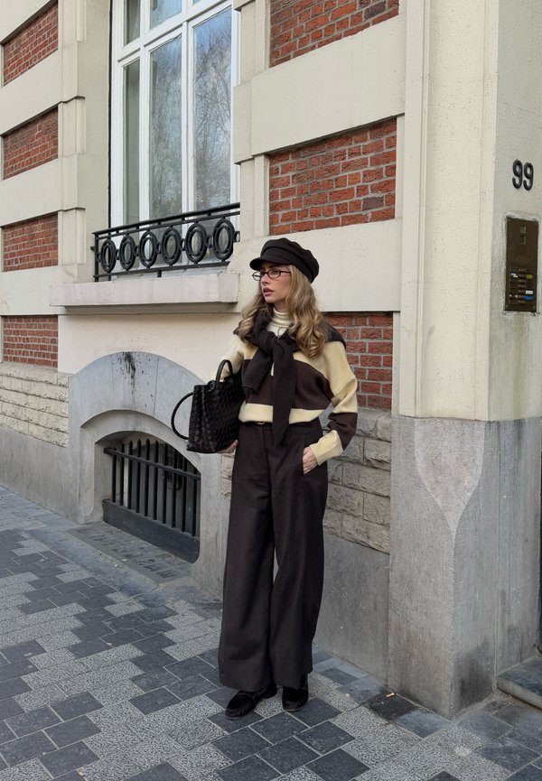 Kvinde iført brun og cremefarvet sweater med vidde bukser, sort hat, briller og sort taske, står på en byfortov nær en murstensbygning.