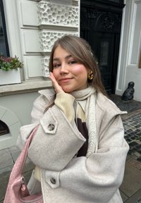 Ljusgrå yllekappa med oversized krage, cremefärgad och brun tröja under, vit stickad halsduk och rosa texturerad handväska. Guldringar framhäver looken.