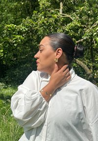 Goldene blumenförmige Ohrringe und ein passendes Armband mit grünen Akzenten. Das Model trägt ein weißes, knöpfbares Hemd mit voluminösen Ärmeln.