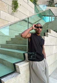 Μαύρο αμάνικο πλεκτό τοπ με υφή, μπεζ πλισέ παντελόνι και μια μικρή μαύρη τσάντα χιαστί, κοντά σε γυάλινες και πέτρινες σκάλες.