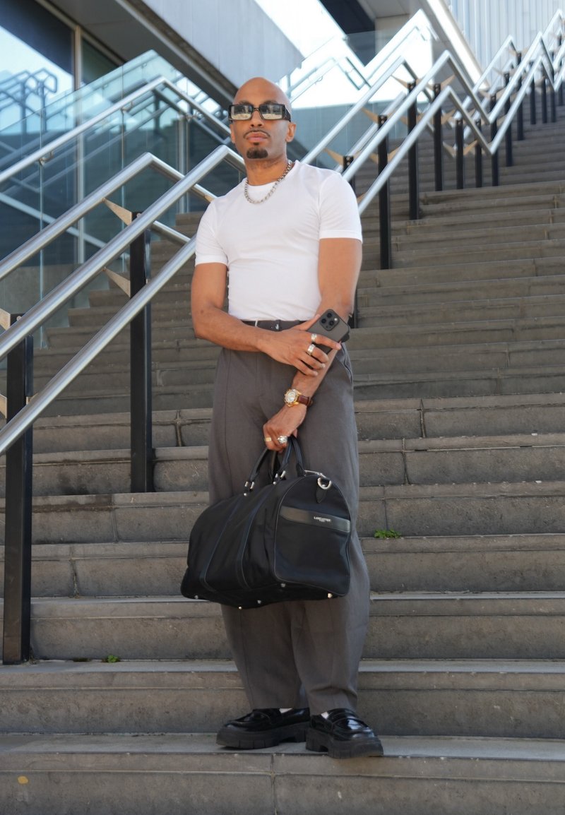 Homme portant un t-shirt blanc et un pantalon gris debout sur des escaliers extérieurs, tenant un sac noir et un smartphone, portant des lunettes de soleil et des bijoux.
