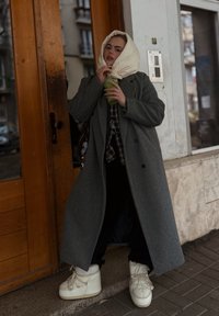 Hosszú szürke kabát kockás ing fölött, fekete nadrág és fehér masszív csizma. Zöld italt tart a kezében, krémszínű sállal a fején.