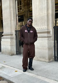 Brun zip-up hoodie med pink stjerne grafik, matchende brune bukser og sorte slouchy støvler. Sat mod en sten søjle baggrund.