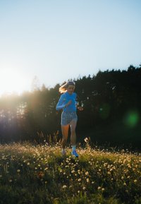 Trkač u svijetloplavoj majici kratkih rukava i šarenenim šortsevima, noseći sunčane naočale, trči kroz polje cvijeća s drvećem u pozadini.