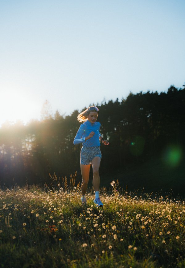 Runner in a light blue short-sleeved top and patterned shorts, wearing sunglasses, running through a field of flowers with trees in the background.