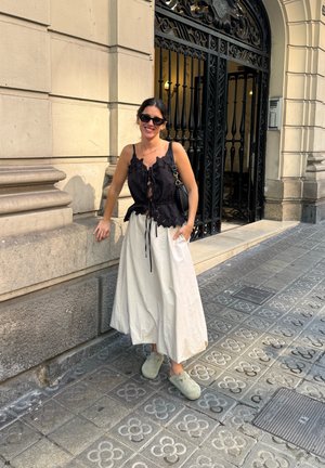 Blusa negra con volantes y lazo ajustable, falda fluida color crema y zuecos verdes, de pie contra una pared de piedra texturizada.