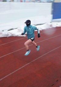 Un coureur portant une veste Adidas turquoise et un short noir avance sur une piste rouge, portant un masque noir et une casquette, avec de la neige en arrière-plan.