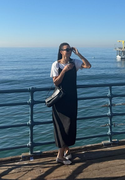 Vestido negro de canalé superpuesto a una camisa blanca, sosteniendo una taza. Lleva gafas de sol y un bolso cruzado negro; unos zapatos de lunares completan el look.