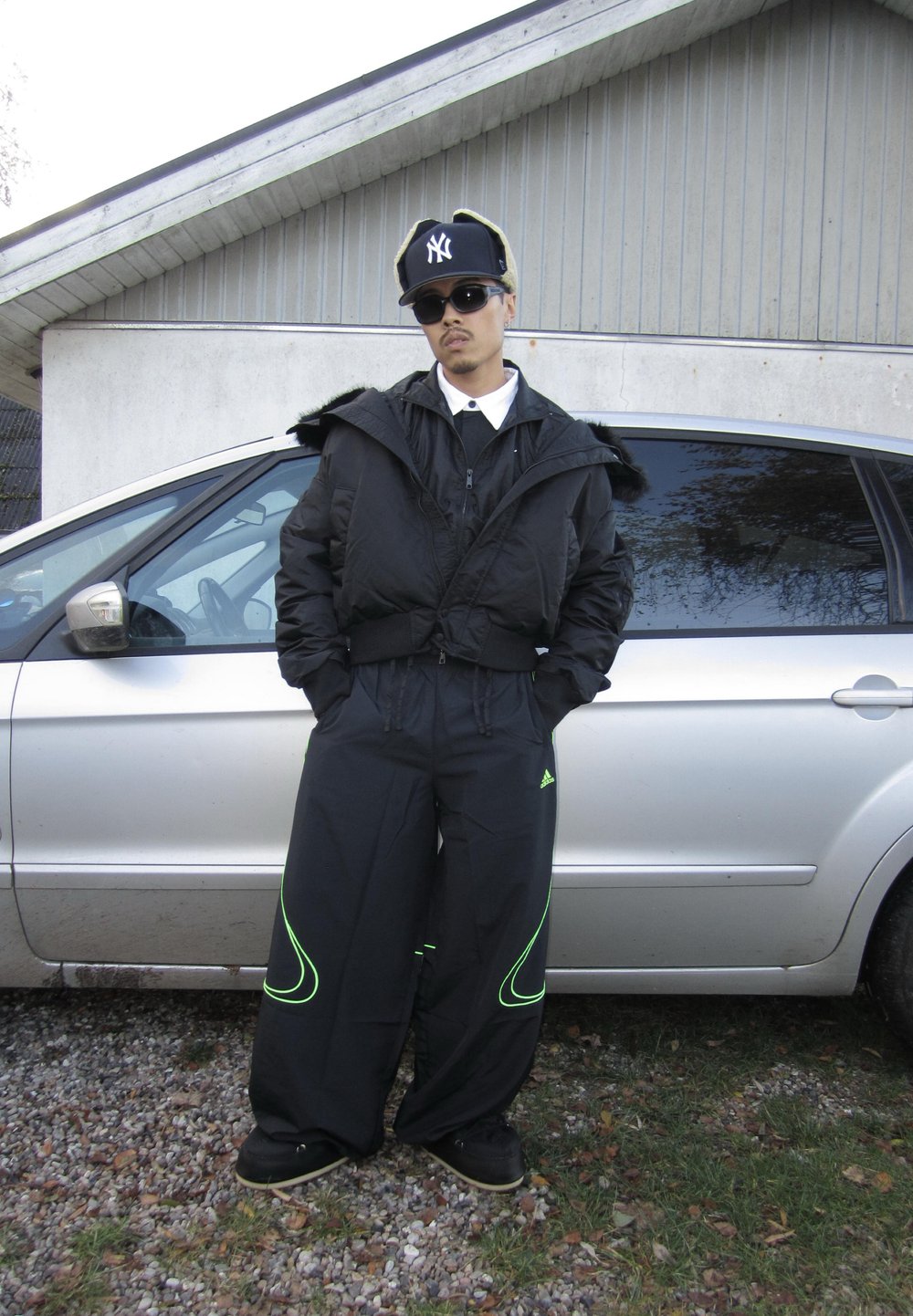 Casaco puff preto com capuz, combinado com calças pretas largas com detalhes verdes. Usado com boné preto e óculos de sol.