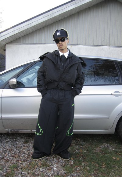 Chaqueta acolchada negra con capucha, combinada con pantalones negros de gran tamaño con detalles verdes. Complementado con una gorra negra y gafas de sol.