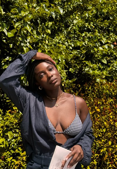 Top corto negro con patrón de cuadros, llevado bajo una camisa de manga larga azul. La modelo sostiene una revista frente a un telón de fondo de follaje verde.