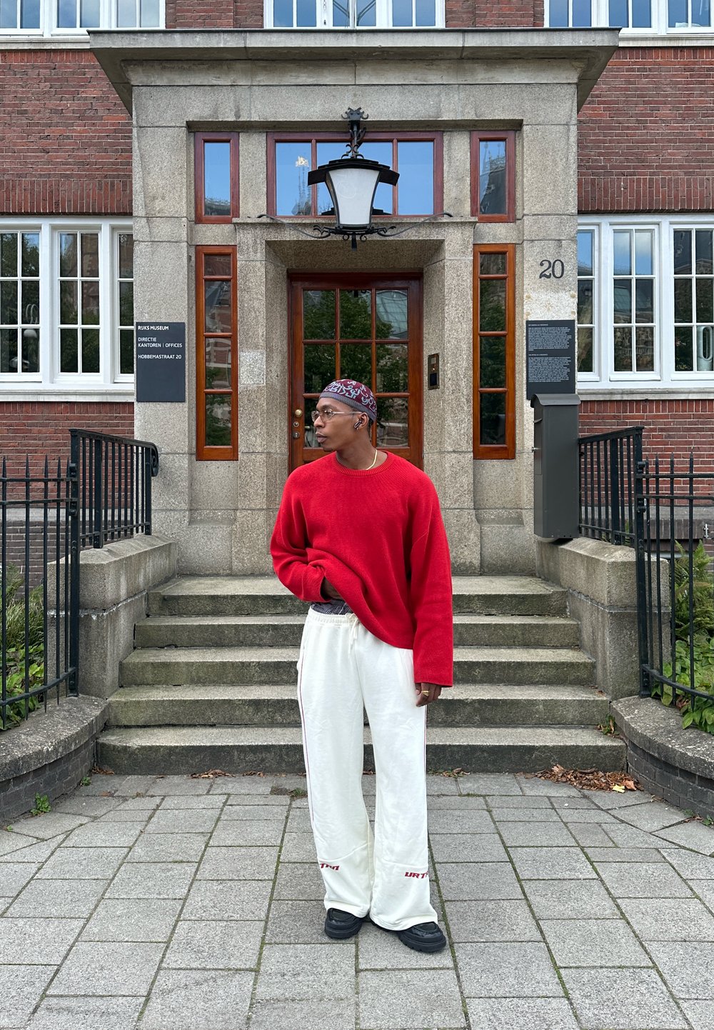 Suéter oversized vermelho, calças de moletom creme com detalhes vermelhos e sapatos pretos. De pé fora de um edifício com degraus de pedra e grandes janelas.