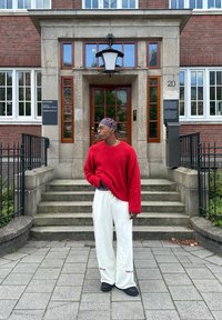Suéter oversized vermelho, calças de moletom creme com detalhes vermelhos e sapatos pretos. De pé fora de um edifício com degraus de pedra e grandes janelas.