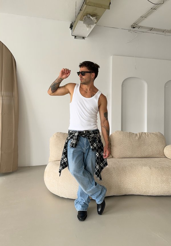 Top branco, óculos de sol pretos, camisa de xadrez amarrada à cintura, jeans azul claro e sapatos pretos. Sofá redondo e bege suave ao fundo.