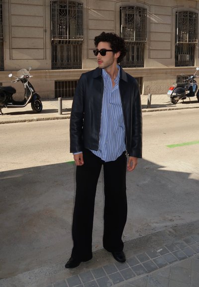 Chaqueta de cuero negra, camisa a rayas azul claro y blanca, pantalones negros de pierna ancha, zapatos negros y gafas de sol; de pie en una calle de la ciudad.