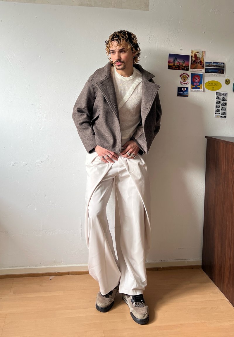 Homme aux cheveux bouclés portant une veste marron, un pull blanc, un pantalon blanc large et des baskets, debout à l'intérieur contre un mur avec des cartes postales.