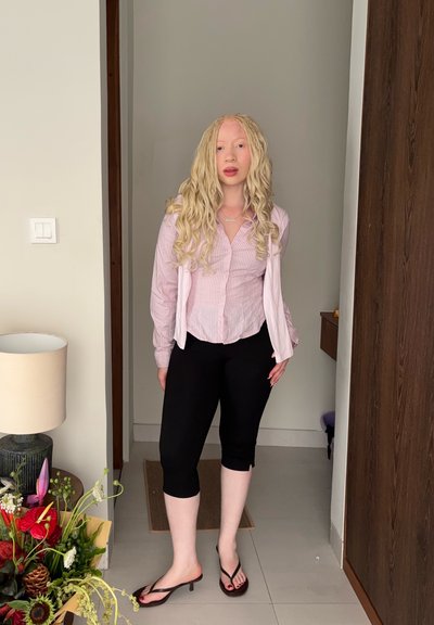 Joven con el cabello rubio rizado y largo, que lleva una camisa rosa a rayas y pantalones negros recortados, de pie en el interior cerca de un arreglo floral y una lámpara.