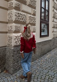 Rode gebreide trui met witte gebreide inzet aan de bovenkant, blauwe jeans met wijde pijpen, bruine laarzen en beige wanten. Lang haar met een rood lint. Achtergrond van een stenen muur.