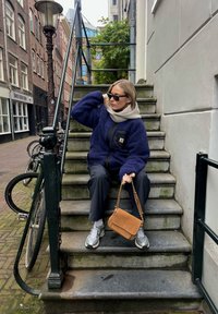 Veste en polaire bleu marine avec accents noirs, écharpe grise, pantalon noir et baskets grises. Sac bandoulière marron. Cadre d'escalier extérieur.