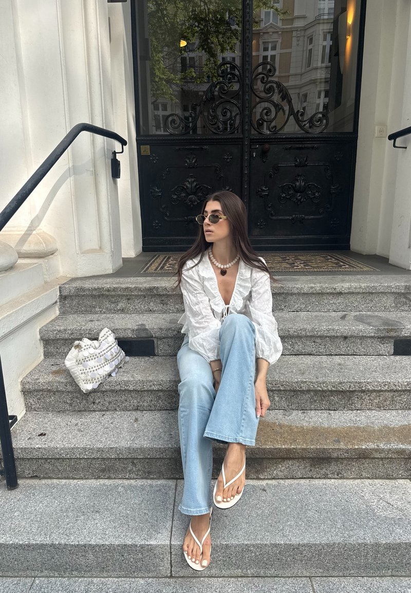 Frau trägt eine weiße Bluse, hellblaue Jeans, Sonnenbrille und weiße Sandalen, sitzt auf Steinstufen mit einer weißen Handtasche neben sich.