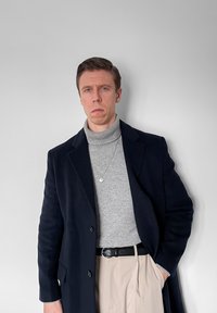 Homme portant un manteau bleu marine, un pull à col roulé gris, un pantalon beige, une ceinture noire et un collier, debout contre un mur gris clair uni.