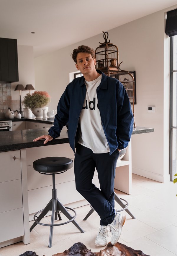 Chaqueta bomber azul marino sobre una camiseta blanca con texto negro, combinada con pantalones azul marino y zapatillas blancas, de pie junto a una encimera de cocina.