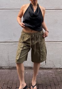 Czarny top na halter, oliwkowe szorty cargo z kieszeniami i czarne klapki na osobie stojącej przed teksturowaną białą ścianą.