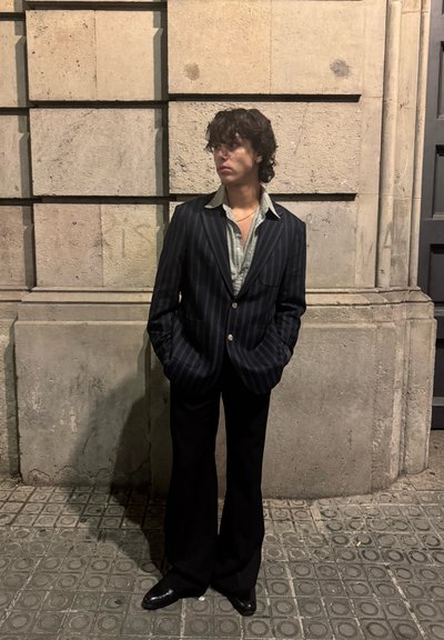 Joven con cabello oscuro y rizado, vestido con un blazer a rayas, camisa con el cuello abierto y pantalones acampanados negros, de pie con las manos en los bolsillos contra una pared de piedra.