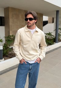 Pullover de fleece bege com colarinho e pala de três botões, combinado com jeans azul claro. O fundo apresenta vegetação e paredes azulejadas.