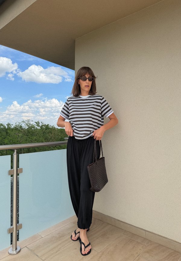 Mujer con gafas de sol negras, camiseta de rayas negras y blancas, pantalones negros holgados y chanclas negras, sosteniendo una bolsa negra tejida en un balcón.