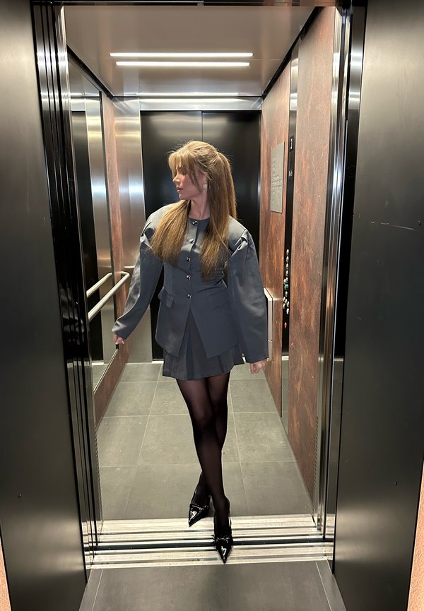 Vrouw in grijze blazer en rok met zwarte panty's en hakken, staand in een moderne lift met spiegels en metalen panelen.