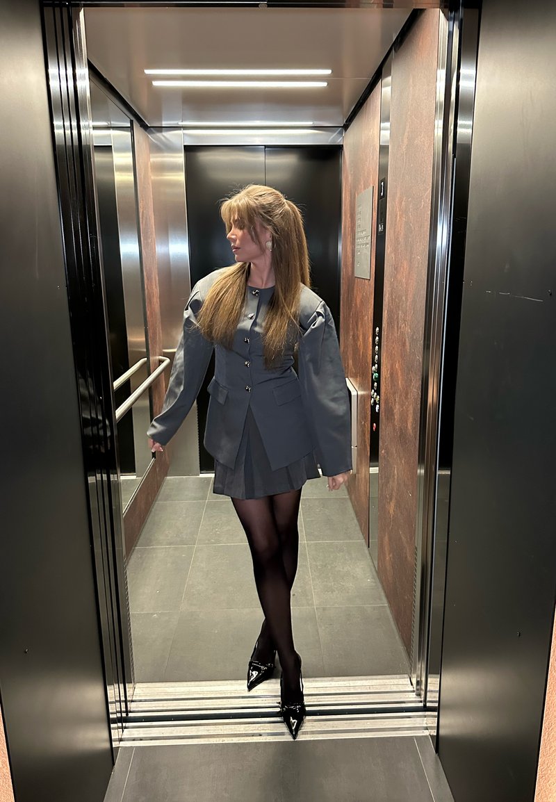 Woman in grey blazer and skirt with black tights and heels standing inside modern lift with mirrors and metal panels.