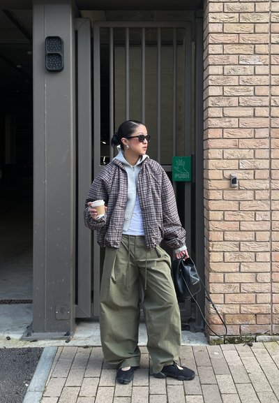 Mujer con gafas de sol que lleva una chaqueta de cuadros y pantalones verdes grandes, sosteniendo una taza de café y un bolso negro cerca de una entrada con puerta y un cartel de "empujar".