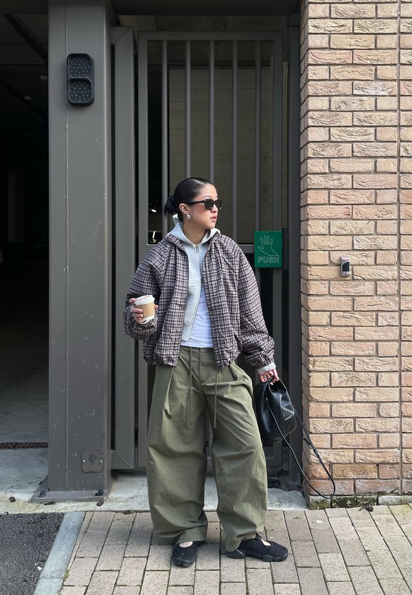 Woman with sunglasses wearing a plaid jacket and oversized green pants holding a coffee cup and black bag near a gated entrance with a push sign.