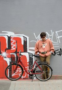 Sarkans velosipēds ar priekšējo grozu, balstīts pret sienu. Cilvēks rozā džemperī ar dizainu, khaki biksēm un saulesbrillēm. Pelēka grafiti fona.