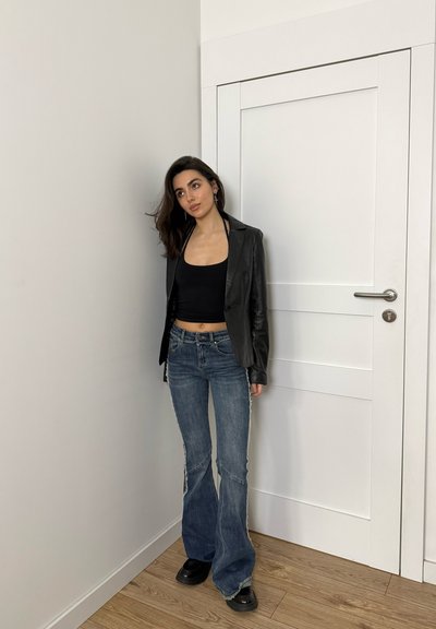 Mujer de cabello oscuro con top corto negro, chaqueta de cuero negra, jeans acampanados azules y zapatos negros, de pie junto a una puerta blanca en el interior.