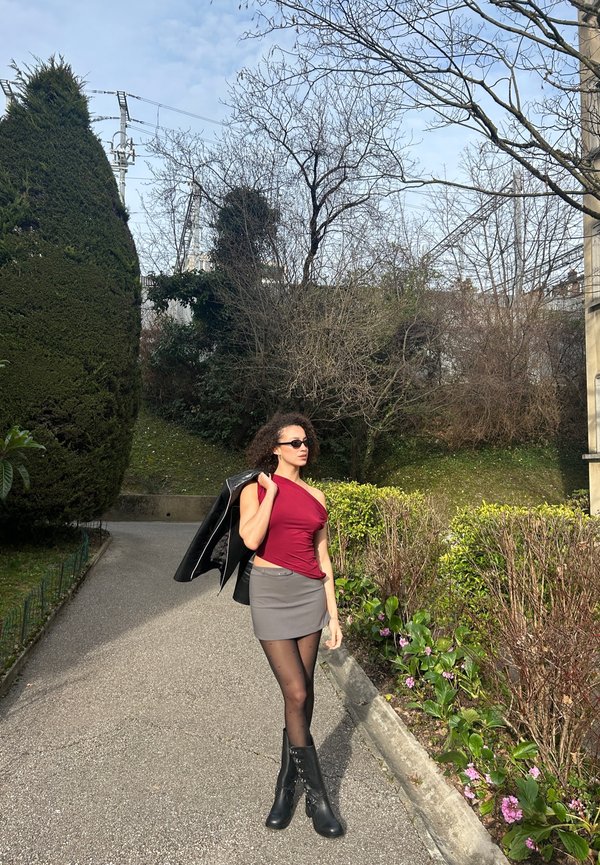Bordo off-shoulder viršus, pilkas mini sijonas, juodos aukštos aulinės botai. Juodas švarkas per petį, permatomos taškuotos pėdkelnės, žaliai lapija fone.