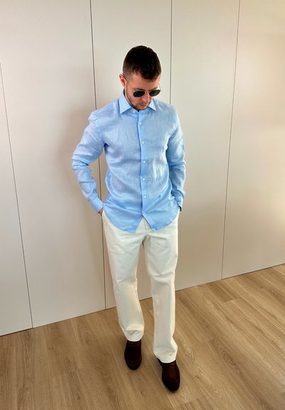 Hombre con camisa azul claro abotonada, pantalones blancos, zapatos oscuros y gafas de sol, de pie en el interior sobre un suelo de madera con las manos en los bolsillos mirando hacia abajo.