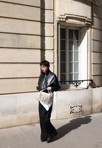 Zwarte leren bomberjack, losse zwarte broek, met een franjes handtas in roomkleur. Verlichte stenen muur en raam op de achtergrond.