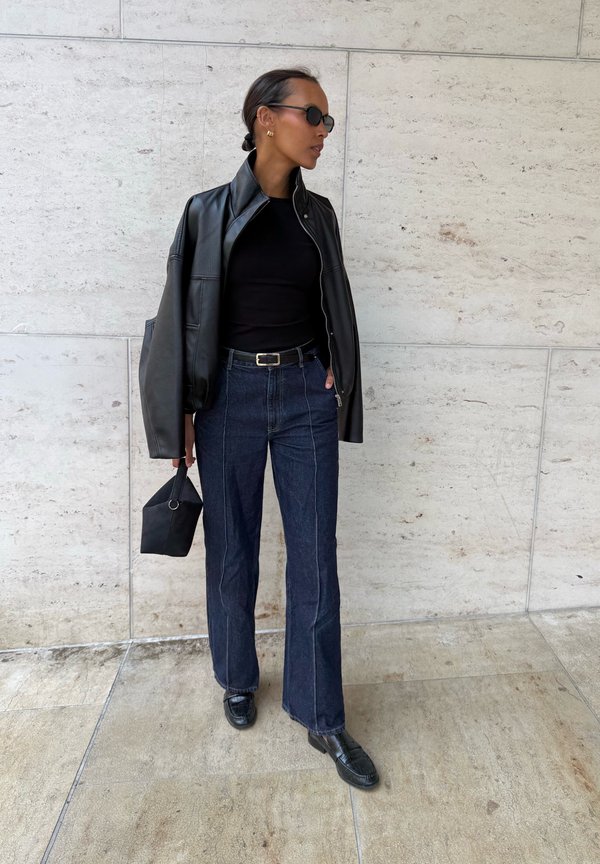 Mujer con chaqueta de cuero negra sobre top negro, vaqueros oscuros de cintura alta, zapatos negros, gafas de sol y sosteniendo un pequeño bolso negro.