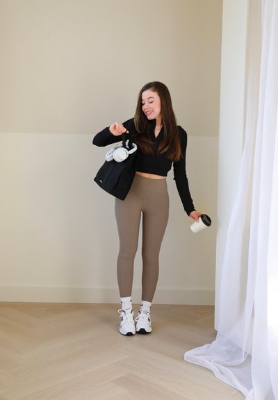 Top negro recortado de manga larga, leggings marrones claros, zapatillas blancas con detalles negros, bolso negro con auriculares blancos y taza de café.