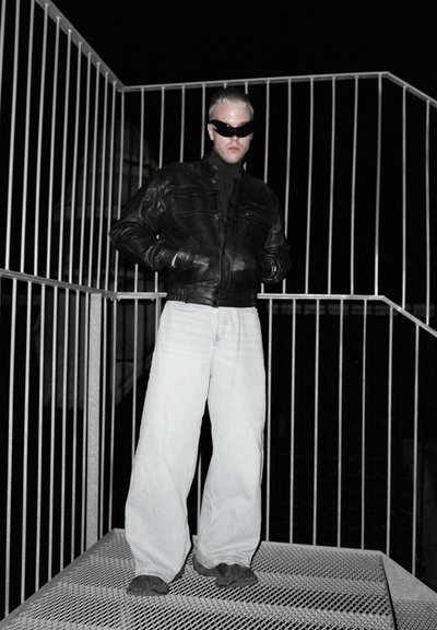 Chaqueta de cuero negra con bolsillos frontales, jeans ligeros de corte holgado en color azul claro, y gafas de sol oscuras. Posicionada sobre una rejilla metálica con barandillas.