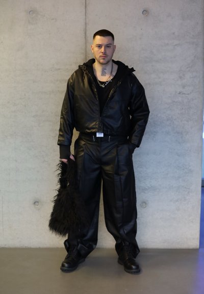 Hombre con pelo corto se encuentra contra una pared de concreto, vistiendo una chaqueta de cuero negra y pantalones, sosteniendo un bolso de piel negra en su mano izquierda.