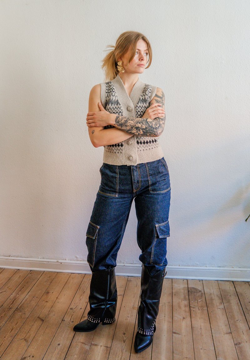 Jeune femme avec un tatouage sur le bras, portant un gilet en tricot à motifs, un pantalon cargo foncé, des bottes noires à clous, debout les bras croisés sur un sol en bois.