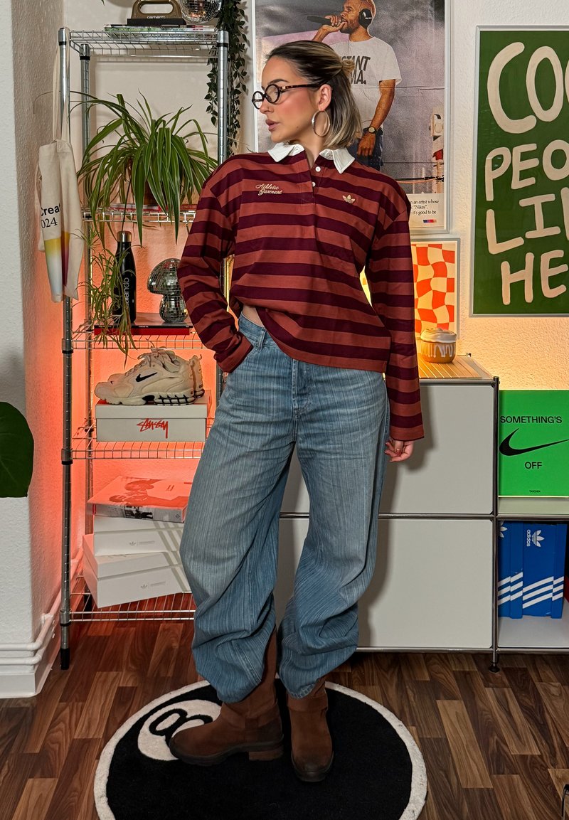 Polo de manga comprida às riscas bordô com gola branca, combinado com jeans azul-claro de pernas largas e botas castanhas até ao tornozelo, em cenário de interior decorado.