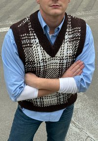 Gilet tricoté à motifs marron et blanc avec un col en V, porté sur une chemise bleu clair ; bras croisés, présentant une finition texturée.