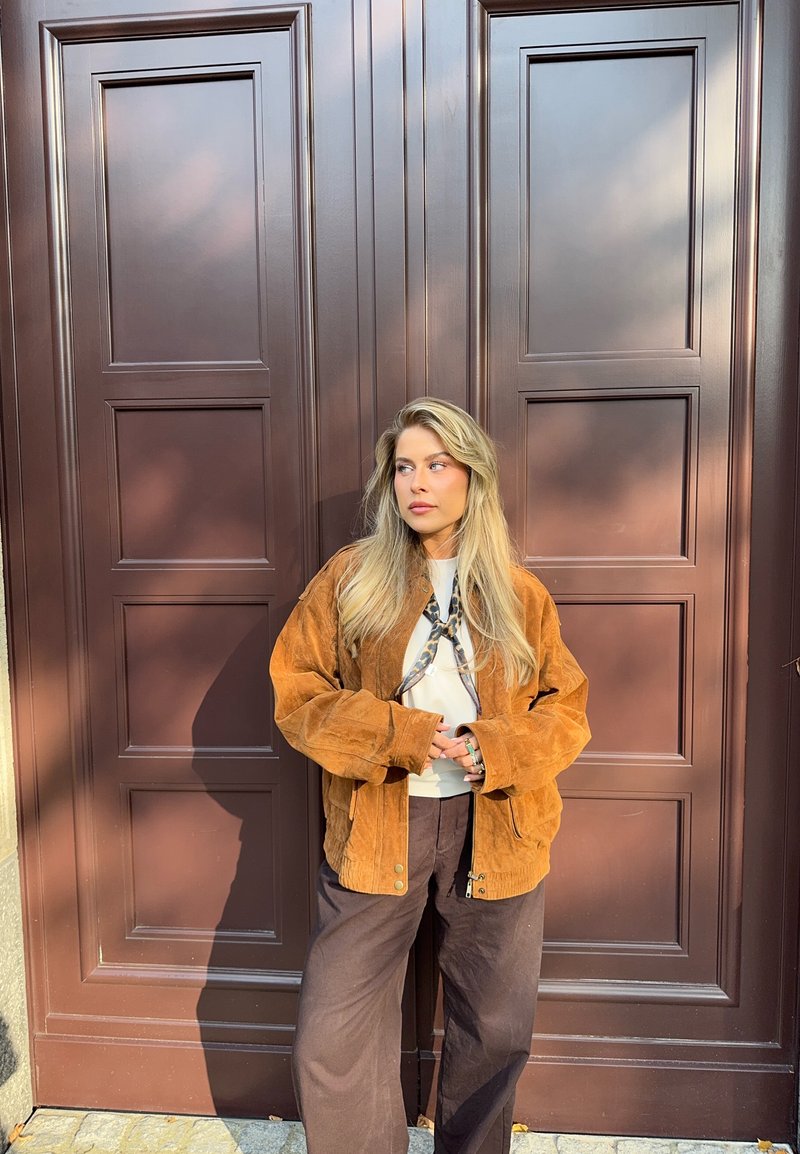 Ruskea corduroy-takki, jossa on rento istuvuus, vetoketjukiinnitys ja taskut, käytetty valkoisen paidan ja tummien leveälahkeisten housujen päällä.