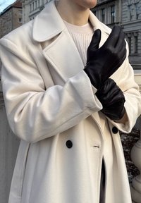 Gants en cuir noir avec des détails de couture subtils, portés par une personne vêtue d'un manteau crème ; la texture lisse contraste avec la douceur du manteau.
