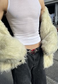 Buste d'une personne portant un débardeur blanc côtelé, une veste duveteuse jaune pâle, un jean noir et une ceinture cloutée avec une boucle en forme de cœur rouge en plein air.